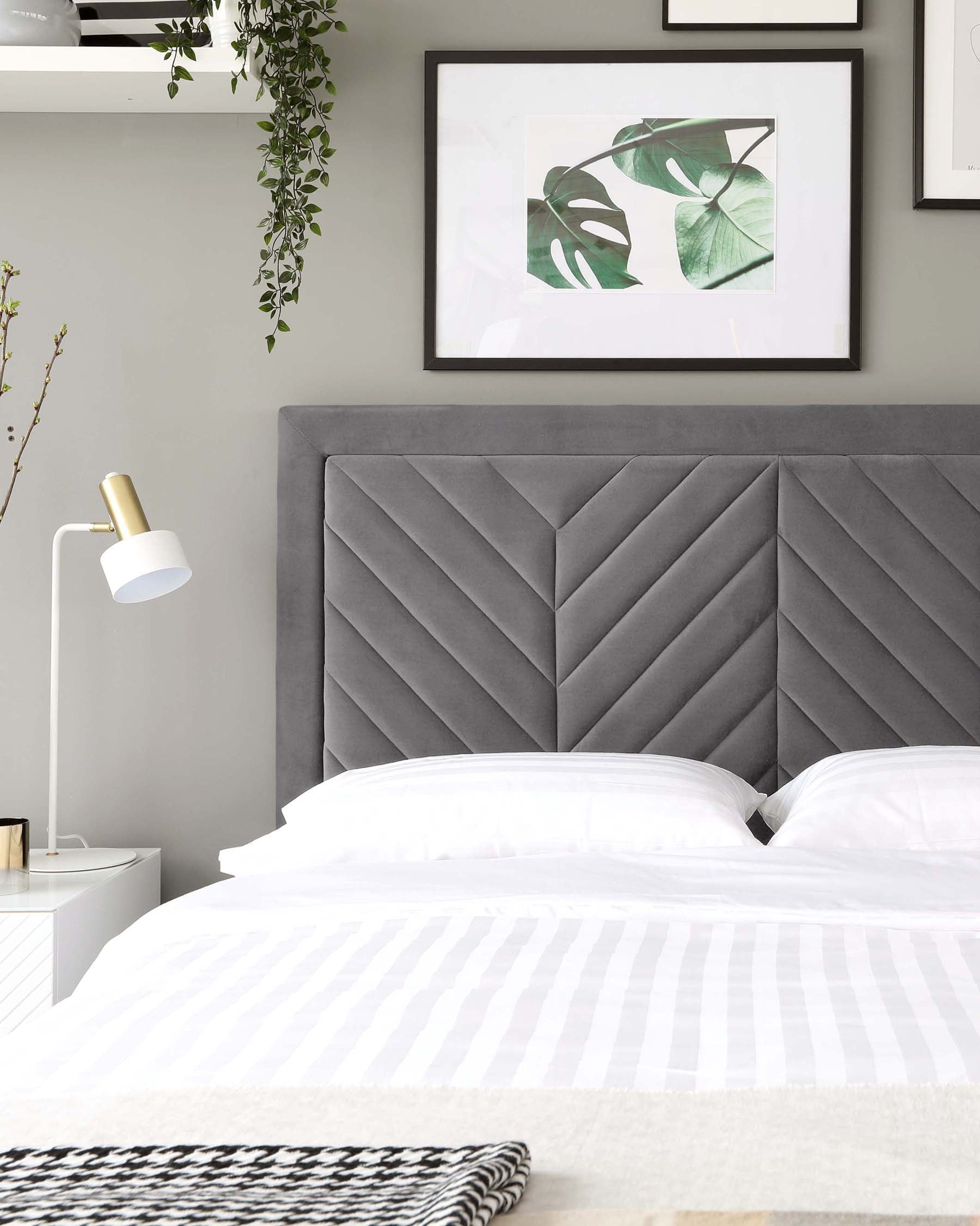 Contemporary bedroom featuring a large upholstered headboard with a geometric tufted design in a plush grey fabric. The bed is dressed in crisp white linens with subtle stripe detailing, complemented by a herringbone throw at the foot of the bed. To the side, there's a minimalist white nightstand with a sleek gold accent on the drawer handle, paired with a modern white and gold table lamp.