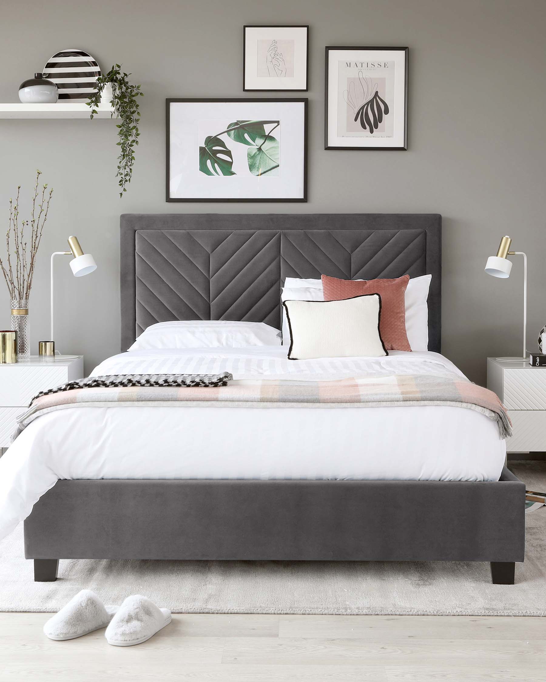 Elegant contemporary bedroom featuring a large grey upholstered bed frame with a high headboard that has geometric tufting. Two white modern bedside tables with gold accents flank the bed, featuring sleek knobs and minimalistic lamps with a white shade and gold base. A soft off-white rug lies beneath the bed, enhancing the room's cosy and stylish aesthetic.