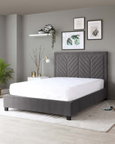 A modern upholstered platform bed with a high headboard featuring a geometric chevron pattern. The bed is dressed in a solid white bedding, and the frame's fabric is a neutral grey, complementing the contemporary bedroom's muted colour palette. Dark wood legs support the bed, and it is set on a light-coloured area rug on a wooden floor, enhancing the minimalist aesthetic of the room.