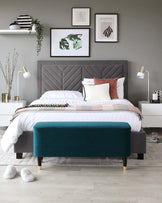 A contemporary styled bedroom showcasing a large tufted headboard in grey fabric, complemented by a matching platform bed frame. At the foot of the bed is an elegant teal upholstered bench with dark wooden legs capped with gold-toned accents. The bed is neatly dressed with a combination of white sheets and a geometric patterned throw. Beside the bed is a small white nightstand with clean lines, supporting a modern white table lamp. The room is anchored by a soft, textured area rug.