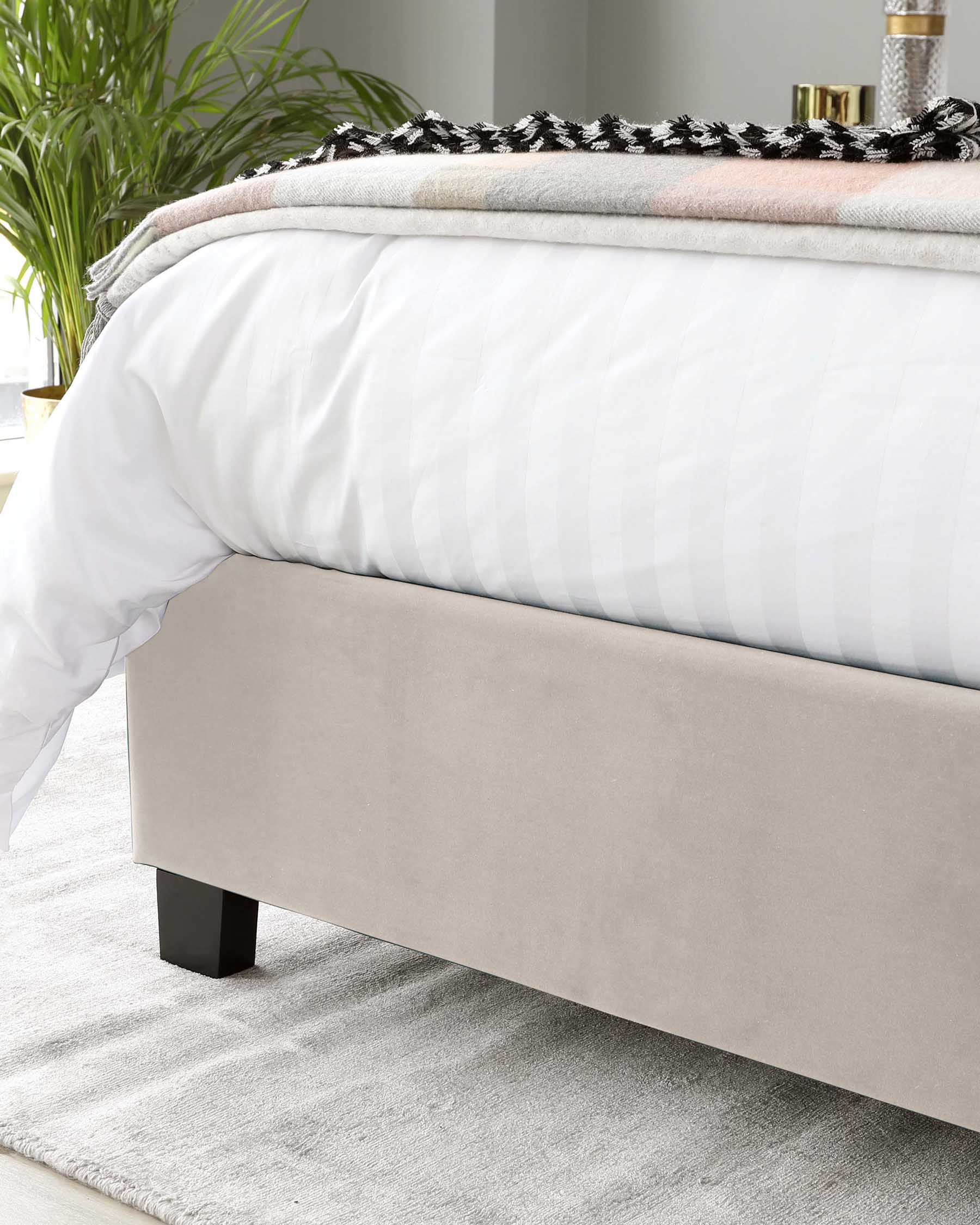 Modern minimalist upholstered bed frame with a beige fabric finish and four black rectangular block legs. Visible is the lower half of the bed with a white duvet, a decorative grey and black throw blanket at the foot, and a plush shag area rug underneath. The bed is staged in a bright room with a hint of greenery from a potted plant in the background.