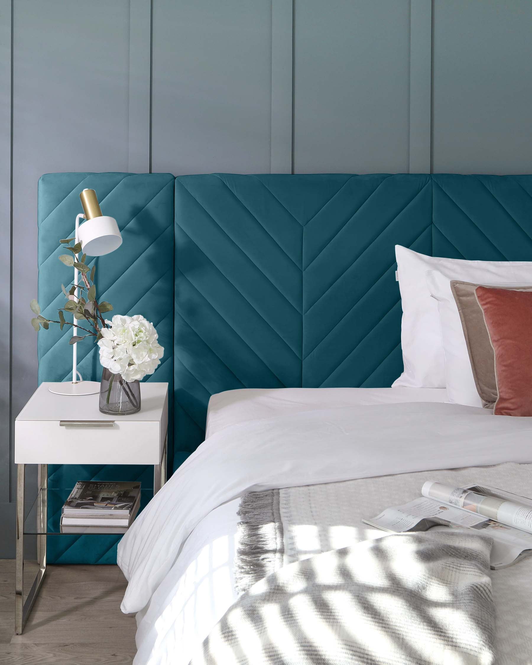 A teal upholstered headboard, sleek white nightstand with a vase of flowers, and stylish bedding create a modern bedroom aesthetic.