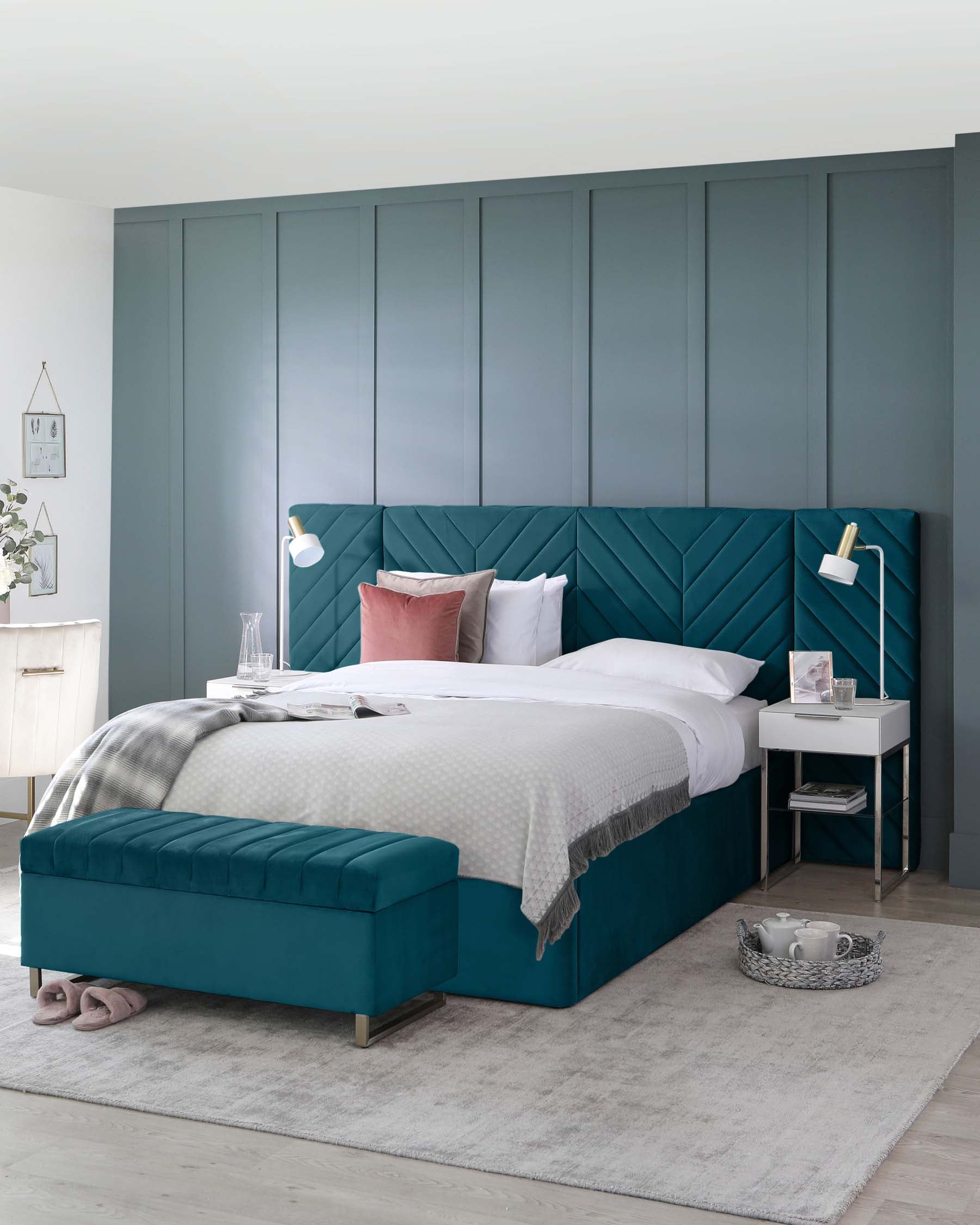 Elegant bedroom furniture consisting of a plush teal upholstered bed with a geometrically patterned headboard and a matching teal ottoman at the foot of the bed. Beside the bed is a white modern nightstand with a lower shelf. The room features a muted grey area rug and wall panelling in a coordinated greyish-blue tone.