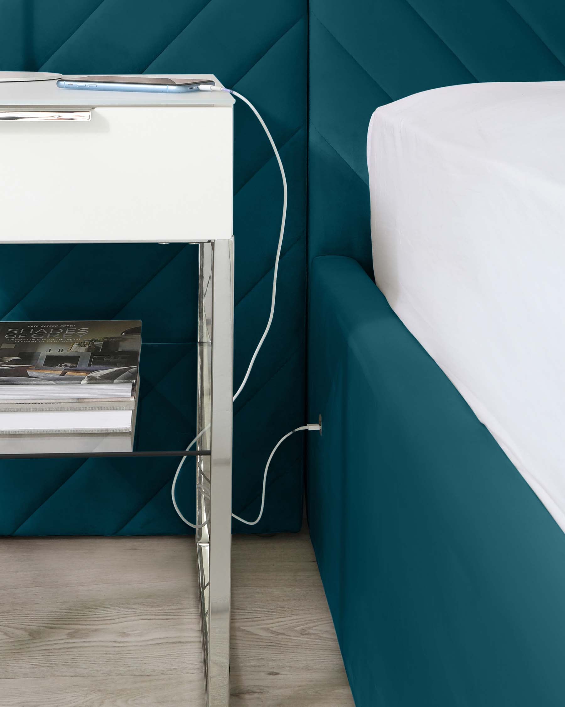 Modern nightstand with white surface, chrome legs, and a lower glass shelf holding magazines, paired with a plush teal bed.