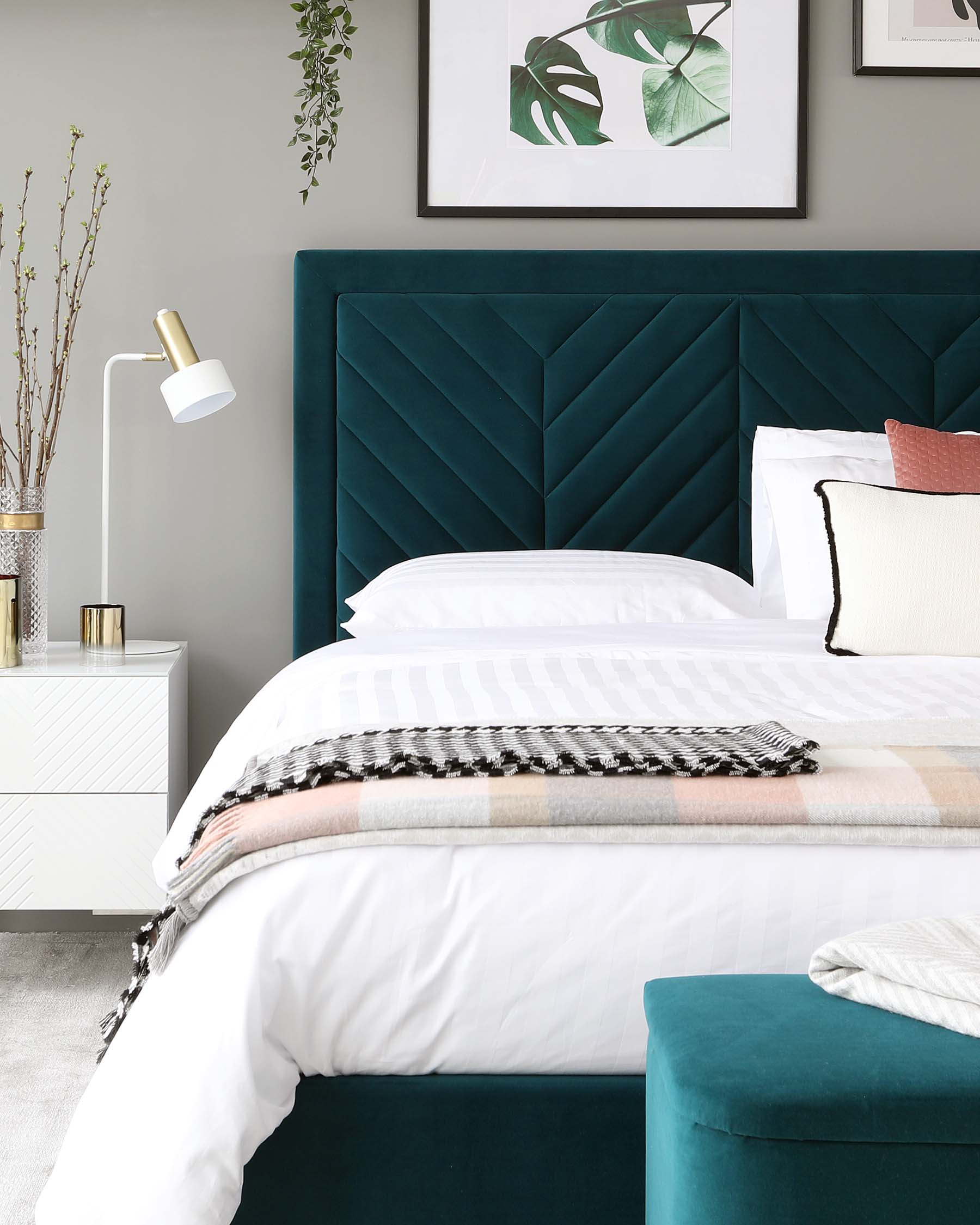 Elegant bedroom setting featuring a large plush emerald green headboard with geometric panel design, accented by crisp white bedding, a white modern side table, and a teal tufted ottoman at the foot of the bed.