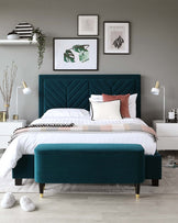 Elegant bedroom featuring a teal upholstered bed with a tufted headboard and a matching teal ottoman at the foot of the bed, both with dark wooden legs capped with gold accents. The bed is dressed in white linens with a multi-coloured striped throw and assorted decorative pillows.