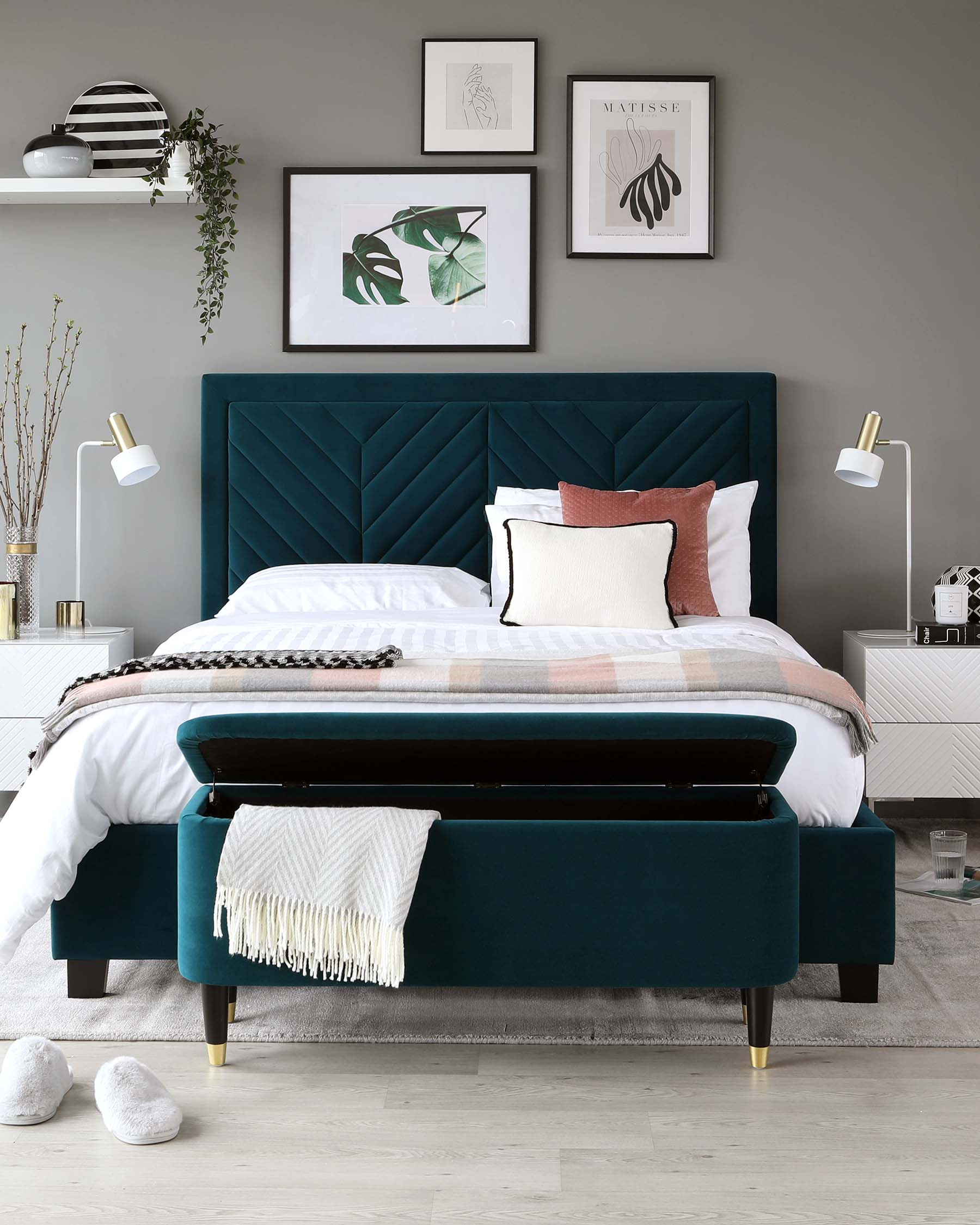 Elegant bedroom featuring a teal upholstered bed with a stylish tufted headboard, complemented by white and pastel bedding with a decorative throw and accent pillows. A matching teal ottoman sits at the foot of the bed. The room is adorned with modern artwork and floating shelves, illuminated by chic wall-mounted lamps.