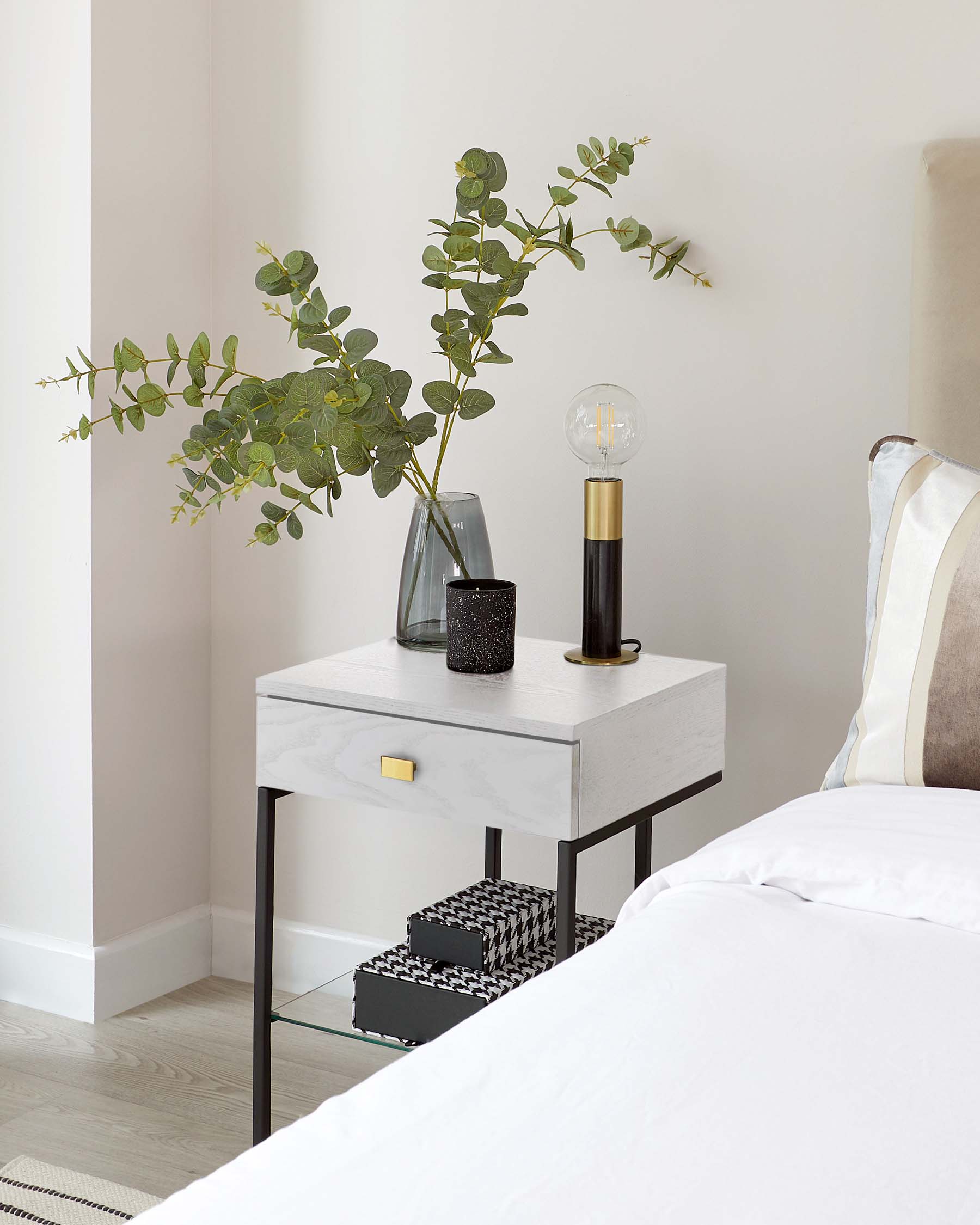 Modern bedside table with a faux marble finish on top, featuring a single drawer with a brass-coloured handle, supported by a minimalist black metal frame and including a lower glass shelf.