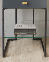 Modern minimalist side table with a matte black metal frame, featuring sleek, straight lines and a luxurious gold handle on the drawer. The table has a clear glass shelf above the drawer and is displayed with a stack of books on the lower shelf. The design is set against a subtle grey wall and on a light wooden flooring, accentuating a contemporary aesthetic.