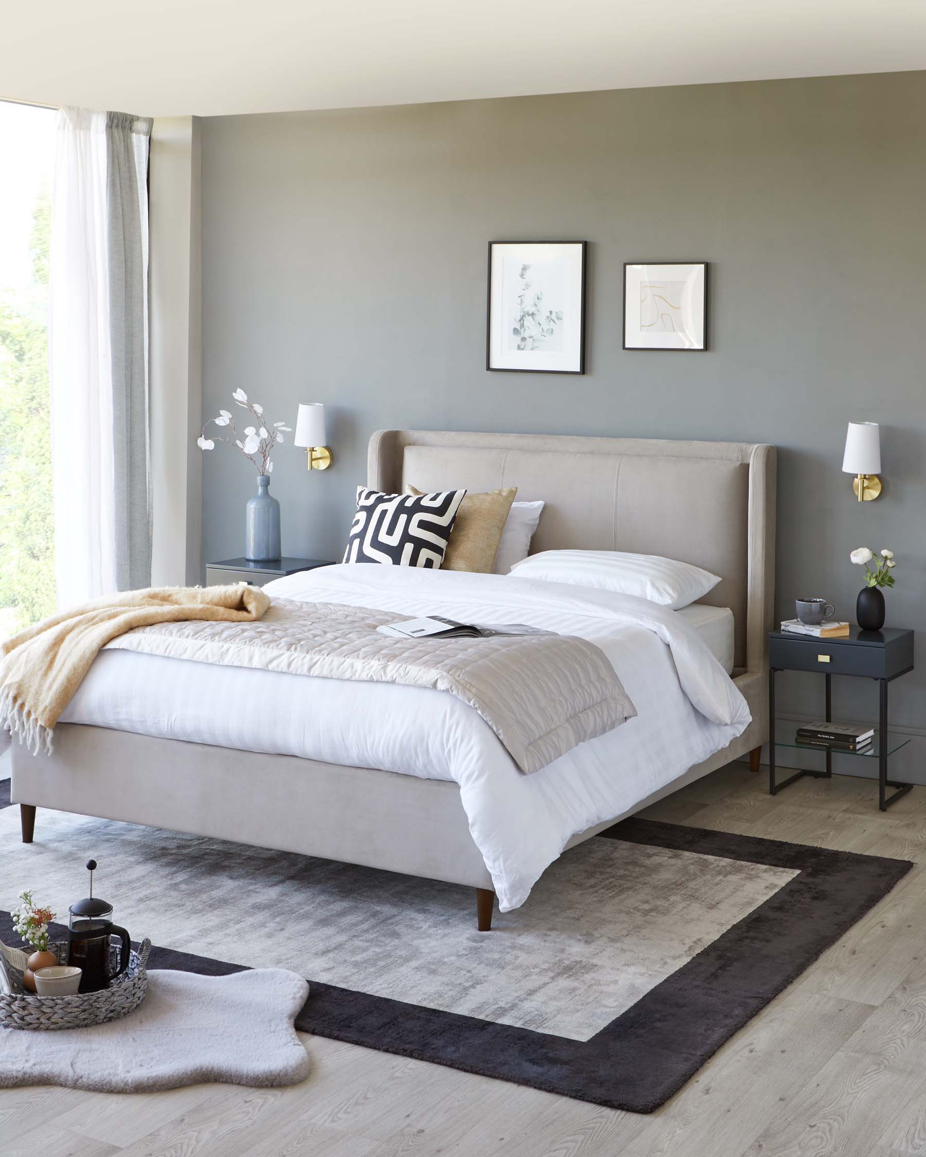 Elegant minimalist bedroom featuring a beige upholstered platform bed with a tall headboard, flanked by two matching dark grey nightstands with drawers, and gold-accented white table lamps. A textured cream throw is laid at the foot of the bed, atop crisp white bedding. On the floor, a contemporary grey area rug with a darker border anchors the space.