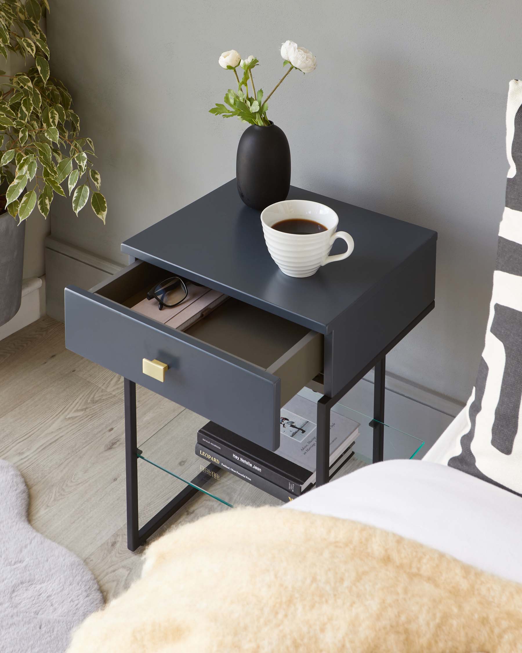 Modern matte charcoal side table with a square top and a pull-out drawer featuring a brass pull handle. The table is supported by a sleek black metal frame with a glass shelf below the drawer for additional storage. On the table's surface is a white ribbed coffee mug and a black vase with white flowers, adding a decorative touch.