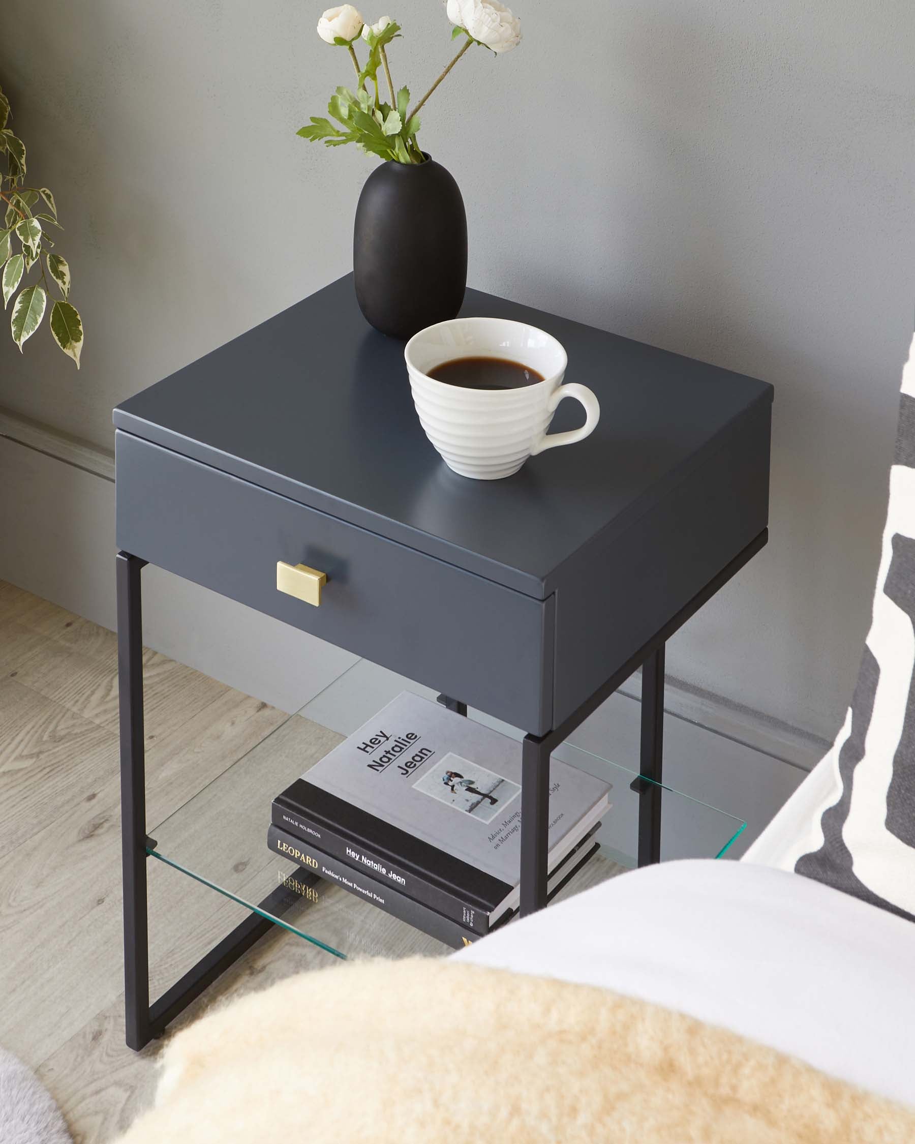 Modern matte charcoal side table with a sleek gold pull on the drawer, featuring a thin black metal frame and a lower glass shelf for additional storage. The table is styled with a white ceramic cup filled with a dark liquid and a matte black vase with white flowers atop, with books placed on the lower shelf.