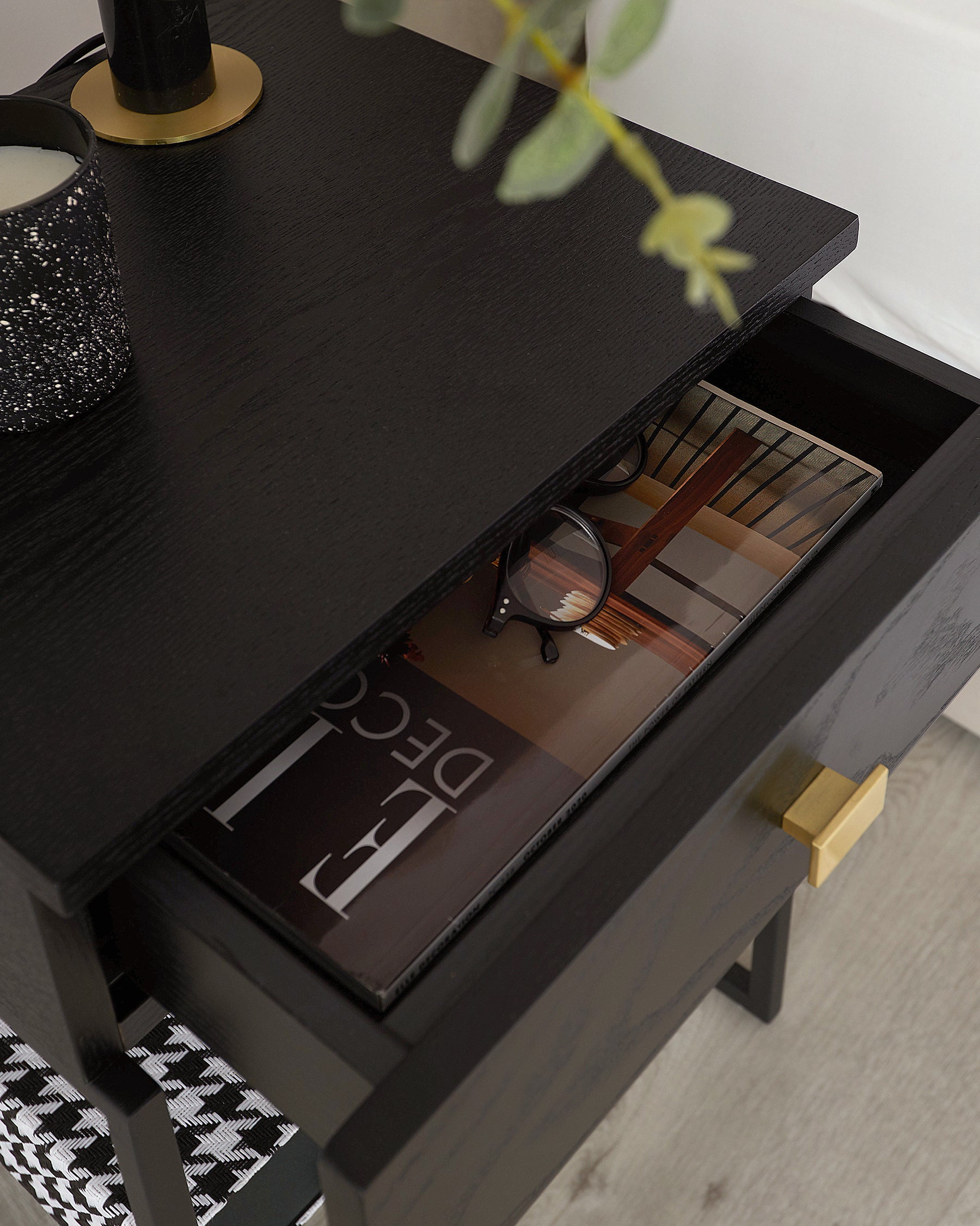 A contemporary black wooden coffee table with a sleek finish and rectangular top, featuring a minimalist design with a partially pulled-out drawer displaying magazines inside. The drawer has a stylish, contrasting gold-coloured metal handle. On the table's surface rests a decorative candle holder and a small potted plant, adding a touch of elegance to the scene.