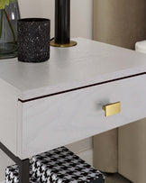 A modern white nightstand featuring a subtle wood grain texture, with a slim dark trim lining the lower edge of the drawer, complemented by a sleek gold-coloured rectangular drawer pull. The nightstand's legs have a contrasting black finish, coordinating with a black and white houndstooth-patterned bench partially visible underneath.