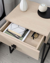 Modern light wood nightstand with an open drawer revealing books and eyeglasses, topped with a white vase and a round candle on a small black tray. The nightstand features a minimalistic design with a metal handle and angular black metal legs.