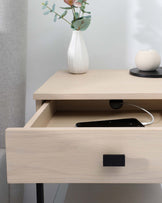 Minimalist light wood nightstand featuring an open shelf and a drawer with a black handle, accessorized with a white vase containing eucalyptus branches and a round, white candle on a black tray. A smartphone is visible inside the partially opened drawer, with a charging cable extending to the top of the nightstand.