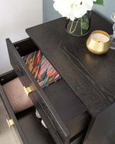 Elegant black wooden sideboard with rich wood grain texture and brass handles, featuring an open drawer with vibrant, colourful lining and a lower shelf storing neatly folded fabrics. The top of the sideboard is adorned with a clear vase of white flowers and a gold-rimmed candle holder.