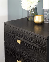 A contemporary black wooden dresser with a textured finish featuring three drawers accented by rectangular, brushed-gold handles. The dresser is accessorized with a clear glass vase holding white flowers, a gold decorative bowl, and a glass candle holder with a white candle.