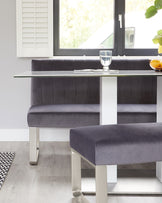 Modern dining furniture featuring a sleek white rectangular table with a clear glass top and polished metal legs, paired with a long, plush bench upholstered in dark grey velvet with coordinating metal legs and vertical stitching on the backrest.