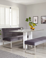 Modern dining room setup featuring a rectangular glass-top table with a white base and sleek, metallic accents. Accompanying the table is a long, tufted, upholstered bench in a plush grey velvet fabric with metallic legs and a matching smaller bench opposite the longer one. The setting presents a contemporary and clean aesthetic.