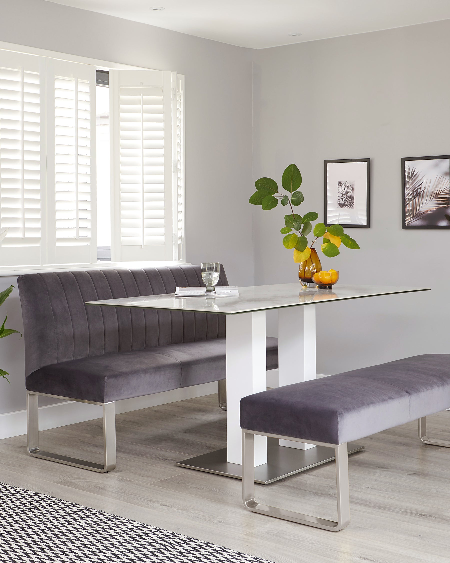 Modern dining room setup featuring a rectangular glass-top table with a white base and sleek, metallic accents. Accompanying the table is a long, tufted, upholstered bench in a plush grey velvet fabric with metallic legs and a matching smaller bench opposite the longer one. The setting presents a contemporary and clean aesthetic.