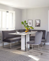 Modern dining set featuring a white rectangular table with metal accents on the base, accompanied by a grey upholstered L-shaped bench and three chairs with curved metal bases and matching grey fabric. A striped area rug lies beneath.