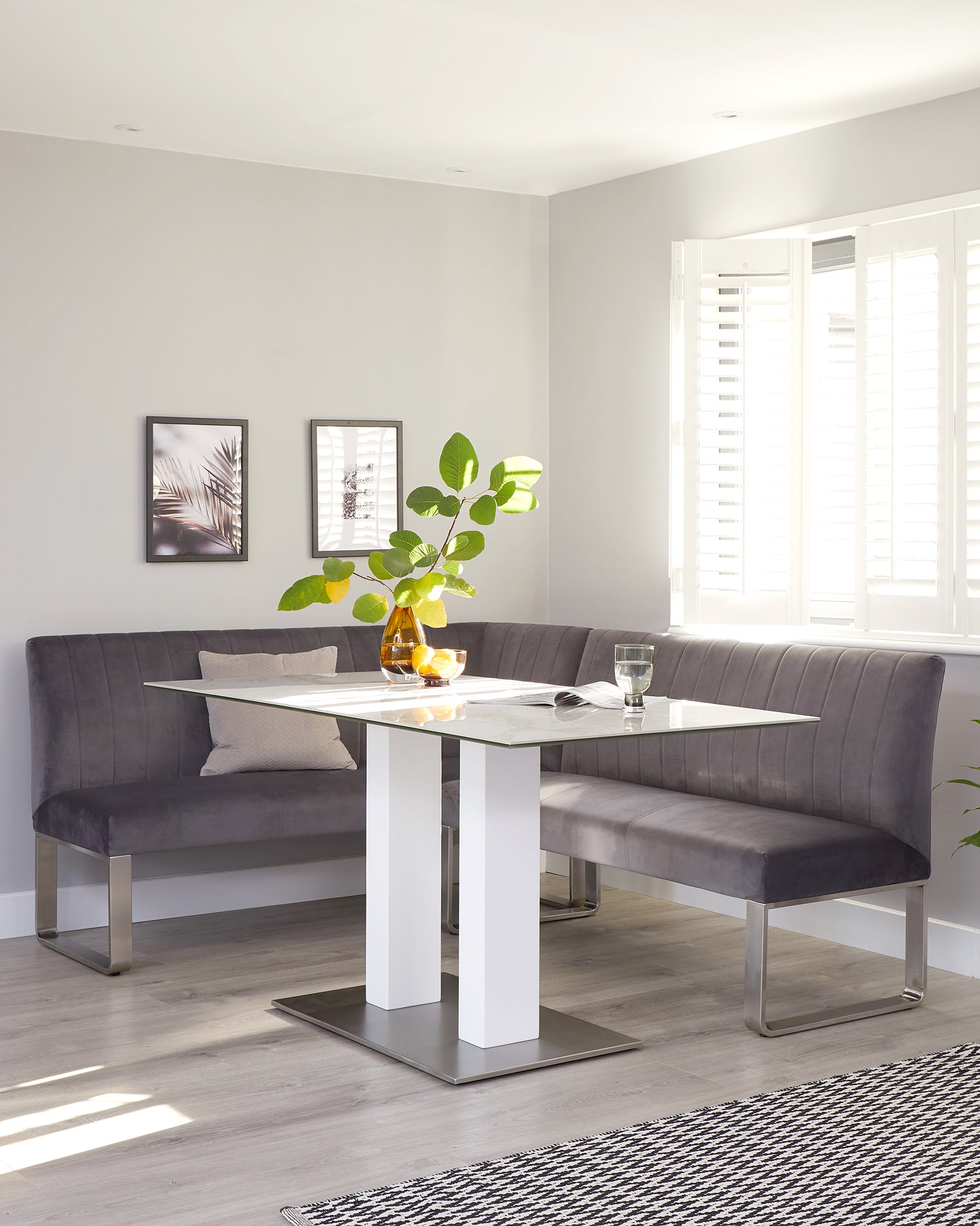 Modern dining set featuring a rectangular glass-top table with a glossy white double pedestal base and brushed stainless steel accents. Accompanied by a curved, tufted back, corner bench upholstered in a plush charcoal fabric with similar stainless steel legs.