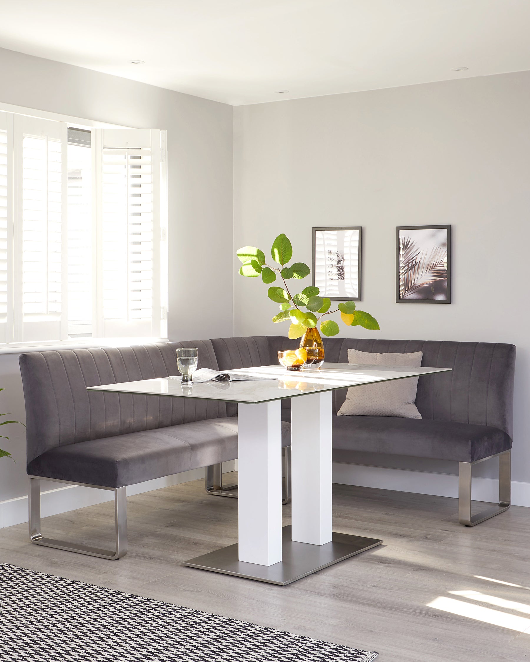 Contemporary dining set featuring a rectangular table with a white marble top, flanked by white high-gloss legs and a chrome-finished metal base frame. Accompanied by an L-shaped corner bench upholstered in grey fabric with horizontal channel stitching, resting on sleek metal legs that mirror the table's design.