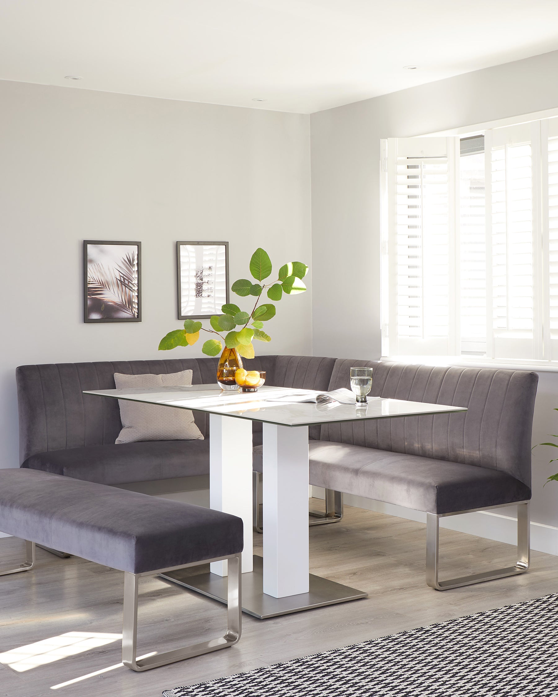 Modern dining set featuring a rectangular glass-top table with a white base paired with a grey upholstered corner bench and a matching backless bench, situated in a well-lit room with neutral tones.