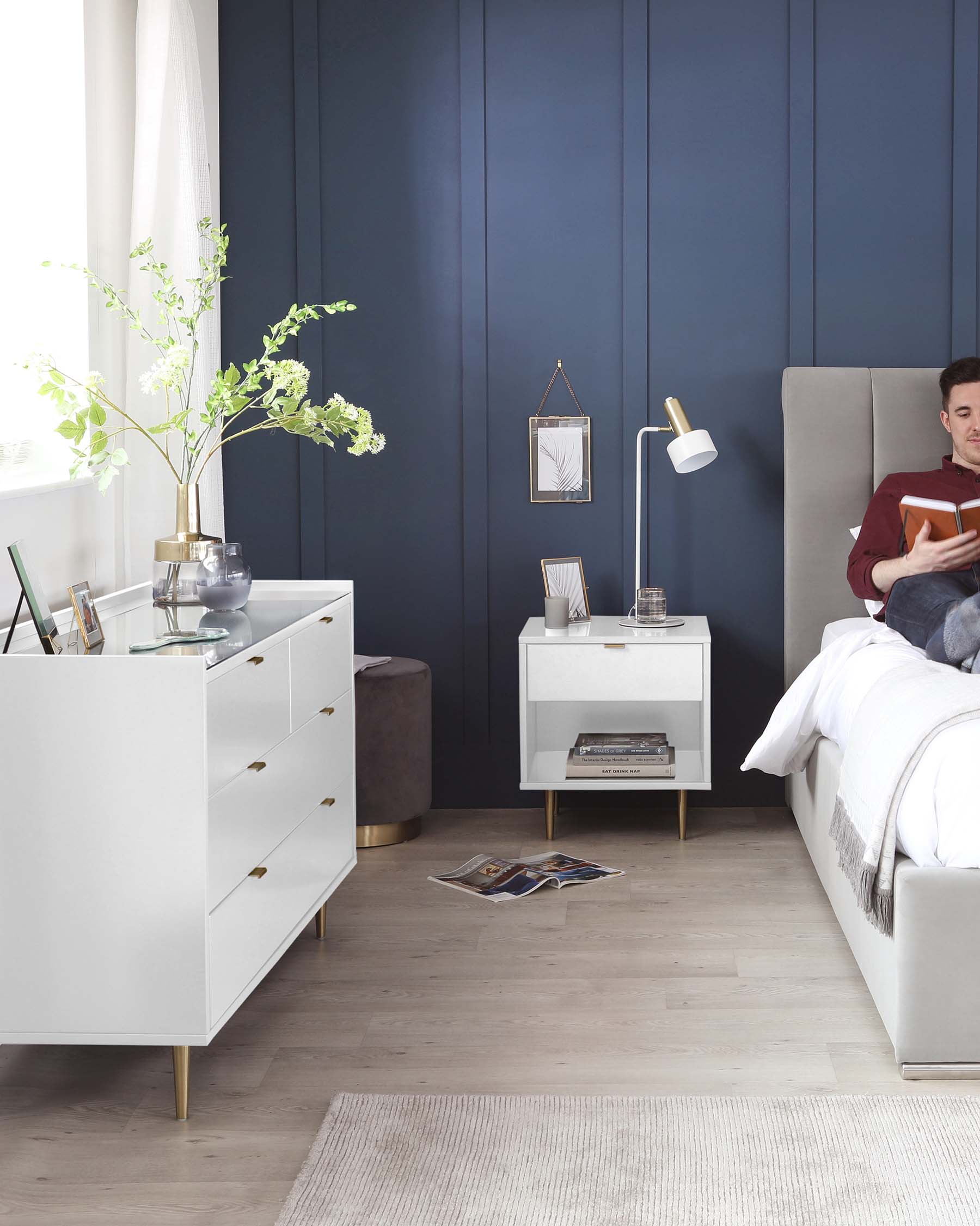 Modern white and gold themed furniture set against a deep blue wall, comprising a tall white dresser with gold handles, a matching white bedside table with a gold base, and a sleek white bed with a high grey upholstered headboard. A coordinating gold floor lamp adds a touch of elegance to the arrangement.