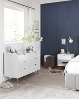 A modern white dresser with gold handles and slender legs positioned against a wall near a window with breezy curtains. Two minimalist framed artworks above a nightstand matching the dresser's style, with a white table lamp and books. A plush, round, grey velvet ottoman is present with golden base detailing. The setting is complemented by light wooden flooring and a soft grey area rug.