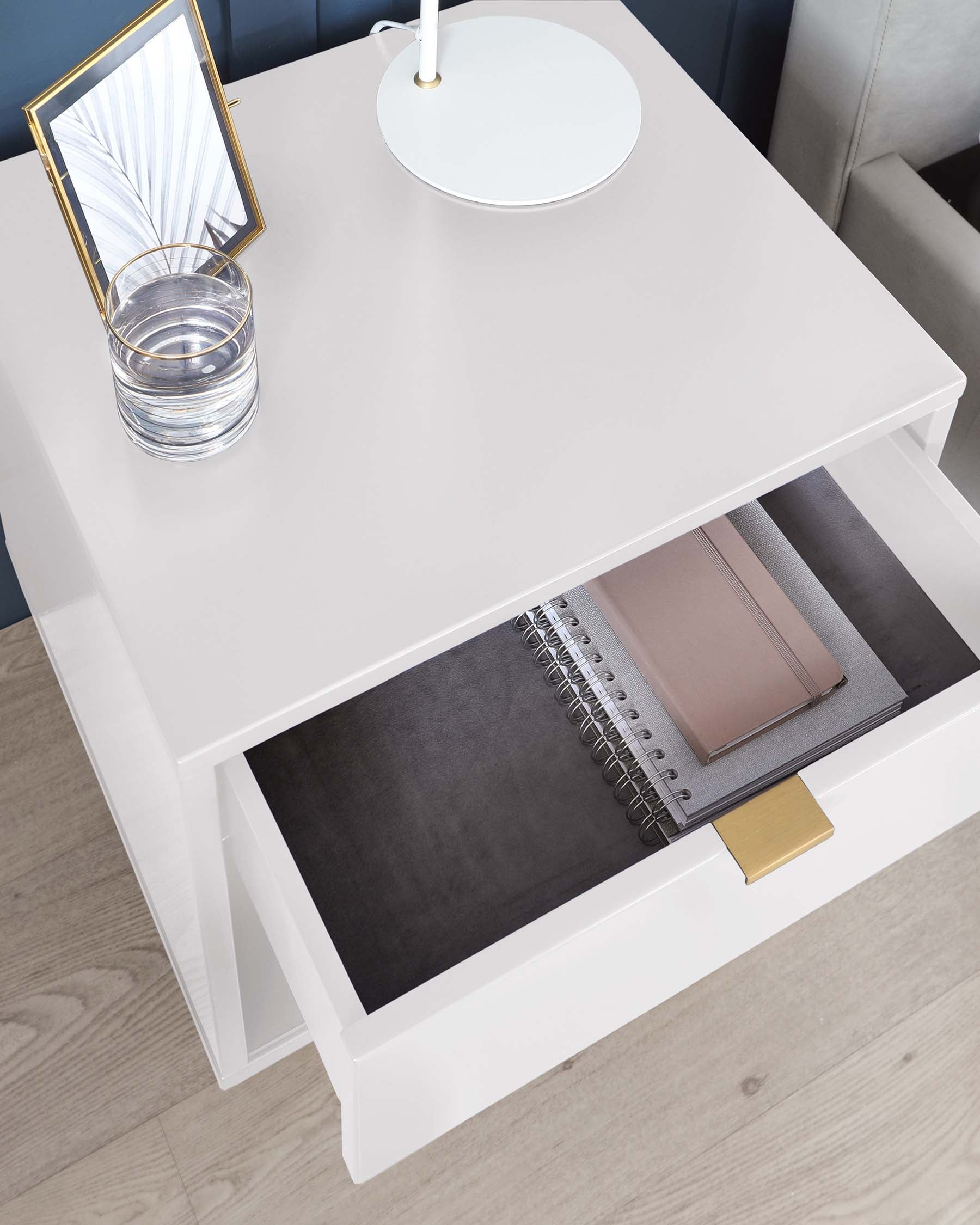 Elegant modern white coffee table with a sleek minimalist design, featuring a functional drawer with a stylish brass pull, partially open to reveal interior storage space with notebooks inside, and the tabletop adorned with a glass of water, a circular white tray, and a framed mirror, all set against a backdrop of a light wooden floor and a portion of a grey sofa.