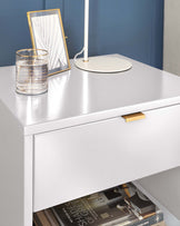 Modern white bedside table with a sleek design, gold-coloured handle and an open lower shelf displaying magazines.