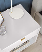 Modern minimalist white side table with a sleek rectangular design, featuring a single drawer with a golden handle and an open lower shelf with magazines. The tabletop is adorned with a round white candleholder and a stack of glass coasters.