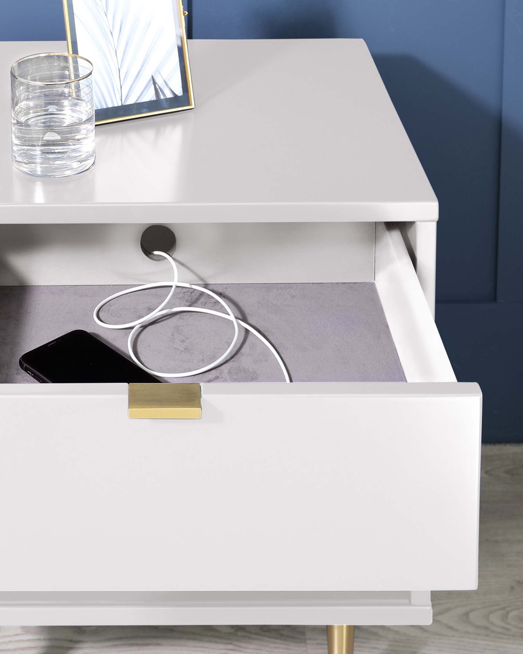 Modern minimalist-style white nightstand with a gold-finished drawer pull, set against a blue wall. The nightstand also features a narrow lower shelf housing a charging cable and a smartphone, partially hidden by the open drawer with grey interior lining. On top, a glass of water and a framed mirror add to the contemporary aesthetic, all above slender golden legs that complement the drawer pull.