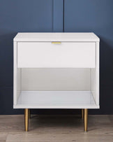 Modern white nightstand with a sleek design, featuring a drawer with a gold handle and an open shelf below, standing on elegant tapered gold legs against a dark blue background.