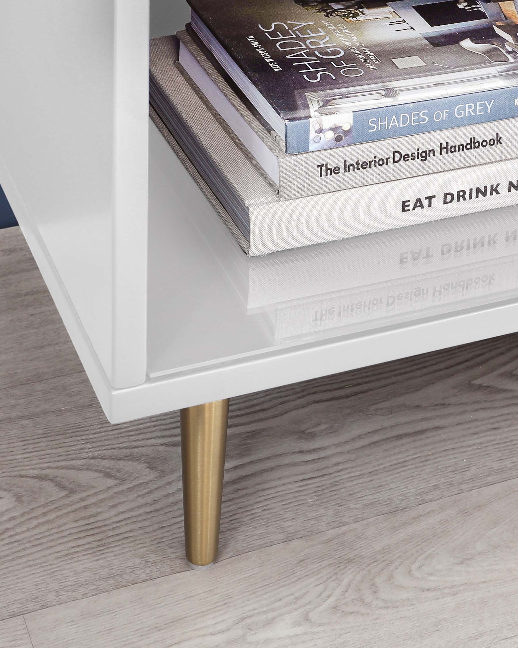 Modern low-profile white coffee table with a glossy finish and an angled metallic gold leg. On its lower shelf, there is a neat stack of hardcover books, illustrating the table's storage functionality.