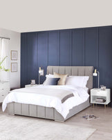 Elegant contemporary bedroom furniture set featuring a king-size bed with an upholstered grey headboard and matching bed frame, crisp white bedding with accent pillows, paired with two white modern nightstands, each with a gold accent lamp, and a sleek white five-drawer dresser. The room has a neutral colour palette with dark blue walls, light grey carpeting, and minimal decorative elements, creating a serene and sophisticated atmosphere.