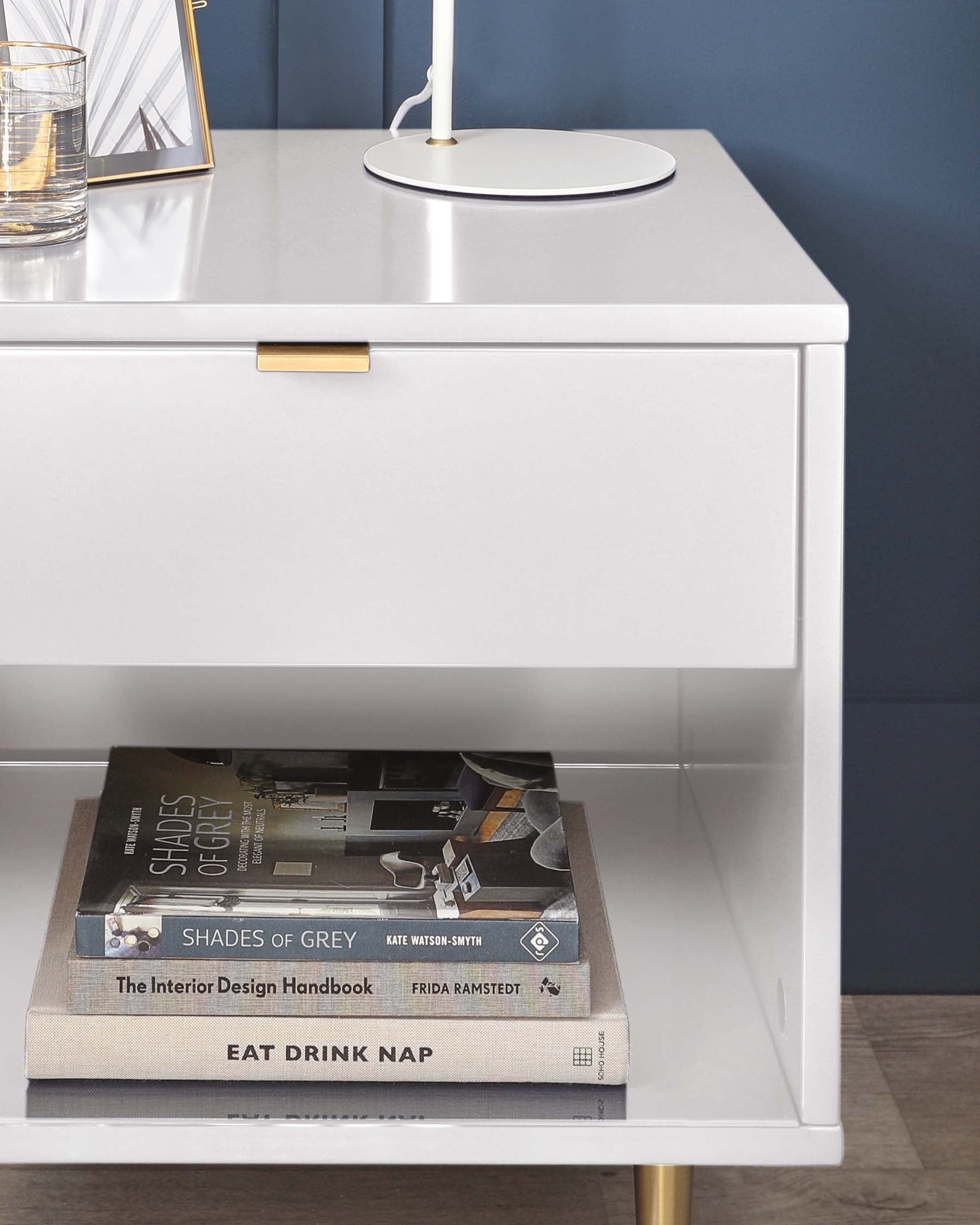 Modern white nightstand with a gold handle on the drawer and gold accented legs. The nightstand features an open lower shelf with two stacked books and the top surface adorned with a white lamp and a decorative glass with a gold frame. The background shows a dark blue wall that contrasts with the furniture's clean and sleek design.