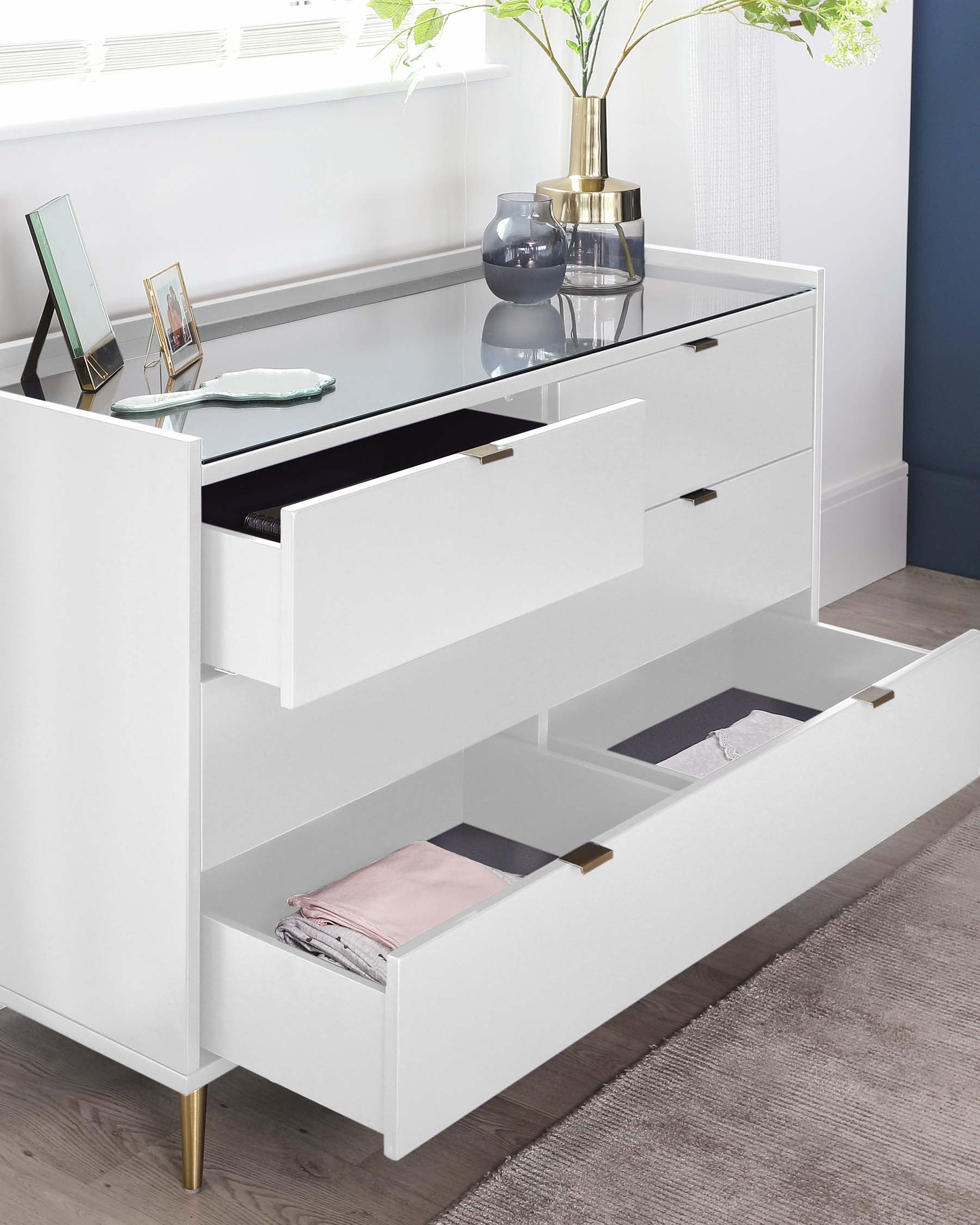 Modern white dresser with a glossy finish, featuring a sleek design, four drawers with two of them partially open displaying folded clothing, accented by brass-finished handles and leg caps, and topped with a clear glass surface hosting decorative items.