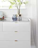 Modern white dresser with clean lines and gold bar pulls on the drawers, topped with decorative items including a blue vase, a metallic lamp, and framed photograph.