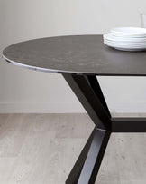 Round table with a dark grey stone finish tabletop and an angular black metal base, displayed with a stack of white plates on top. The table is set against a light hardwood floor and a neutral wall background.