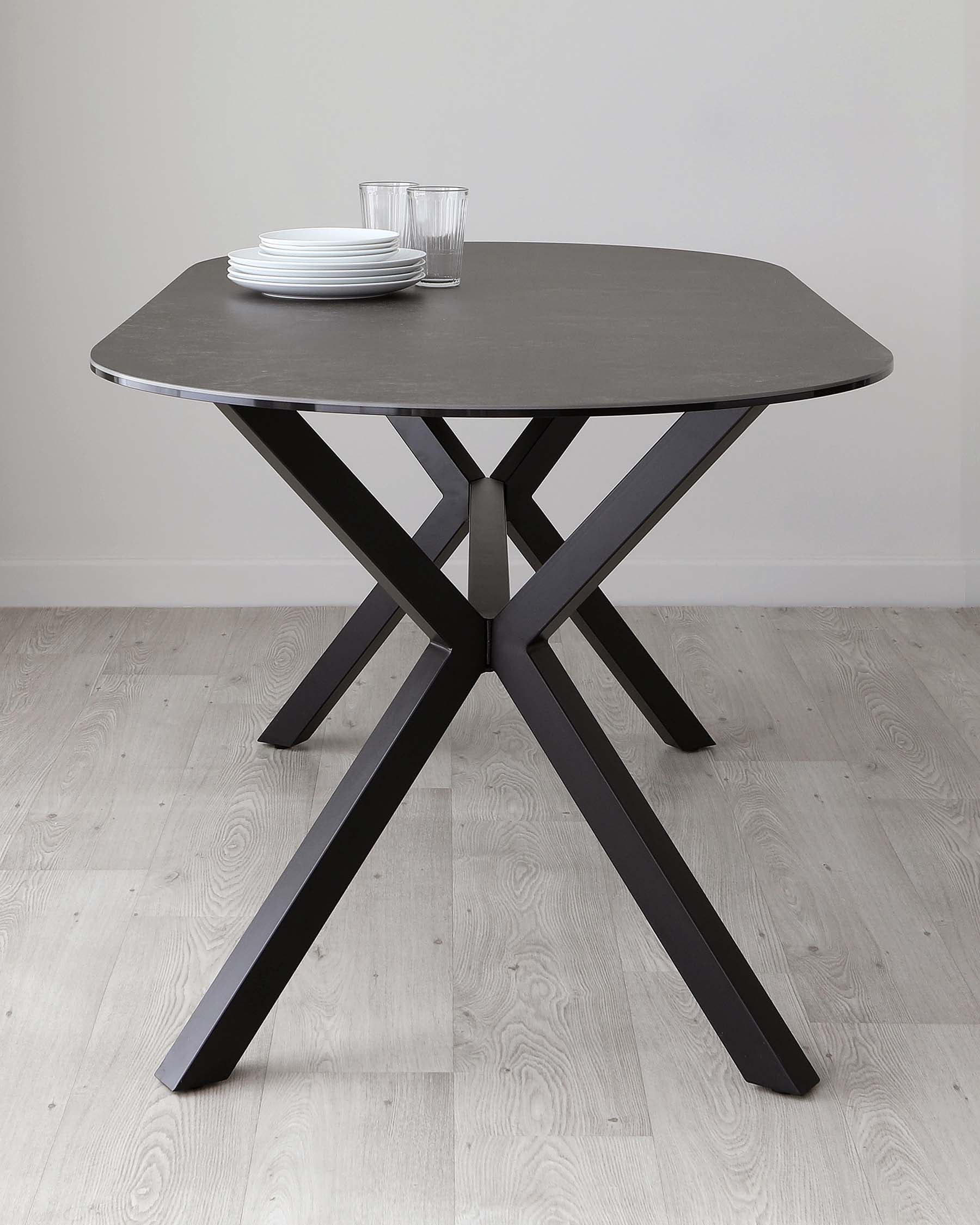 Modern round dining table with a matte black tabletop and unique crisscrossed black wooden legs, set on a light wooden floor. Additional items include a stack of white plates and two clear glasses atop the table.