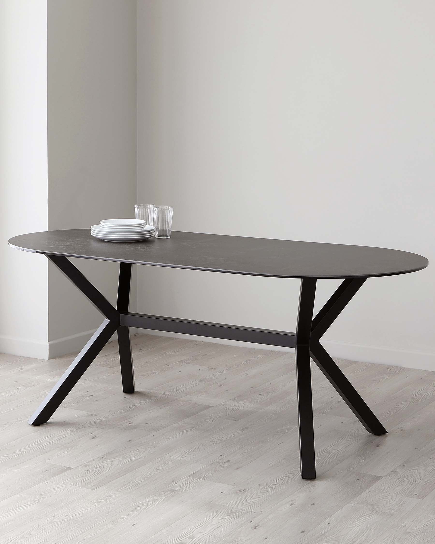 A modern oval dining table with a dark stone finish top and a striking black metal base that features an X-shaped design, set against a neutral backdrop with a stack of white plates and clear glasses on top.