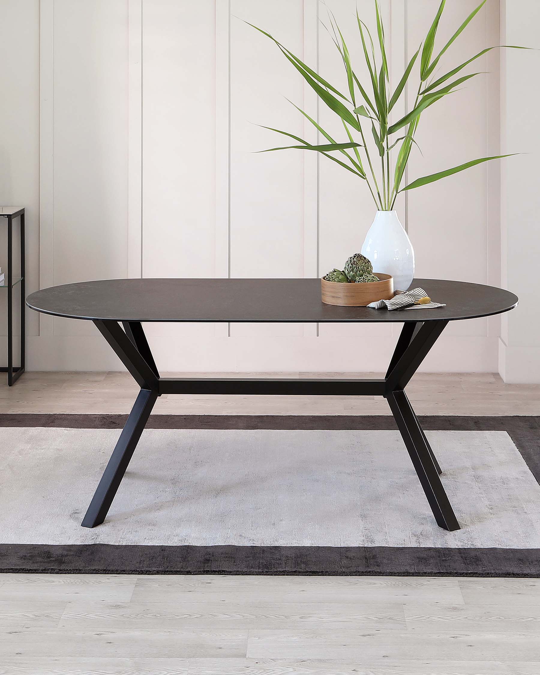 Modern oval-shaped dining table with a dark, espresso finish top and contrasting black, angular metal legs positioned on a light-grey area rug. The tabletop is accessorized minimally with a round wooden tray holding succulents, a seashell, and a tall white vase with green foliage.