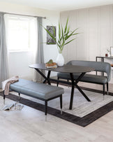 Modern dining room set featuring an oval dark grey table with a matte finish and angled black legs, paired with two grey upholstered seats with slender metallic legs and a matching grey upholstered bench. The set is arranged on a gradient grey area rug over light hardwood flooring.