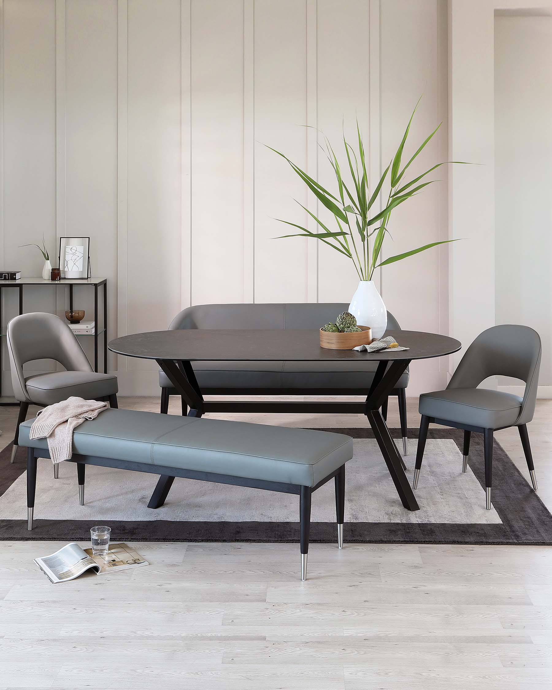 Modern dining room featuring an oval-shaped dark wood table, accompanied by two matching sleek chairs with curved backs and a padded leather bench. The pieces are set on a textured grey area rug over light hardwood flooring. A minimalist black shelving unit accessorized with small decorative items is also present in the background.