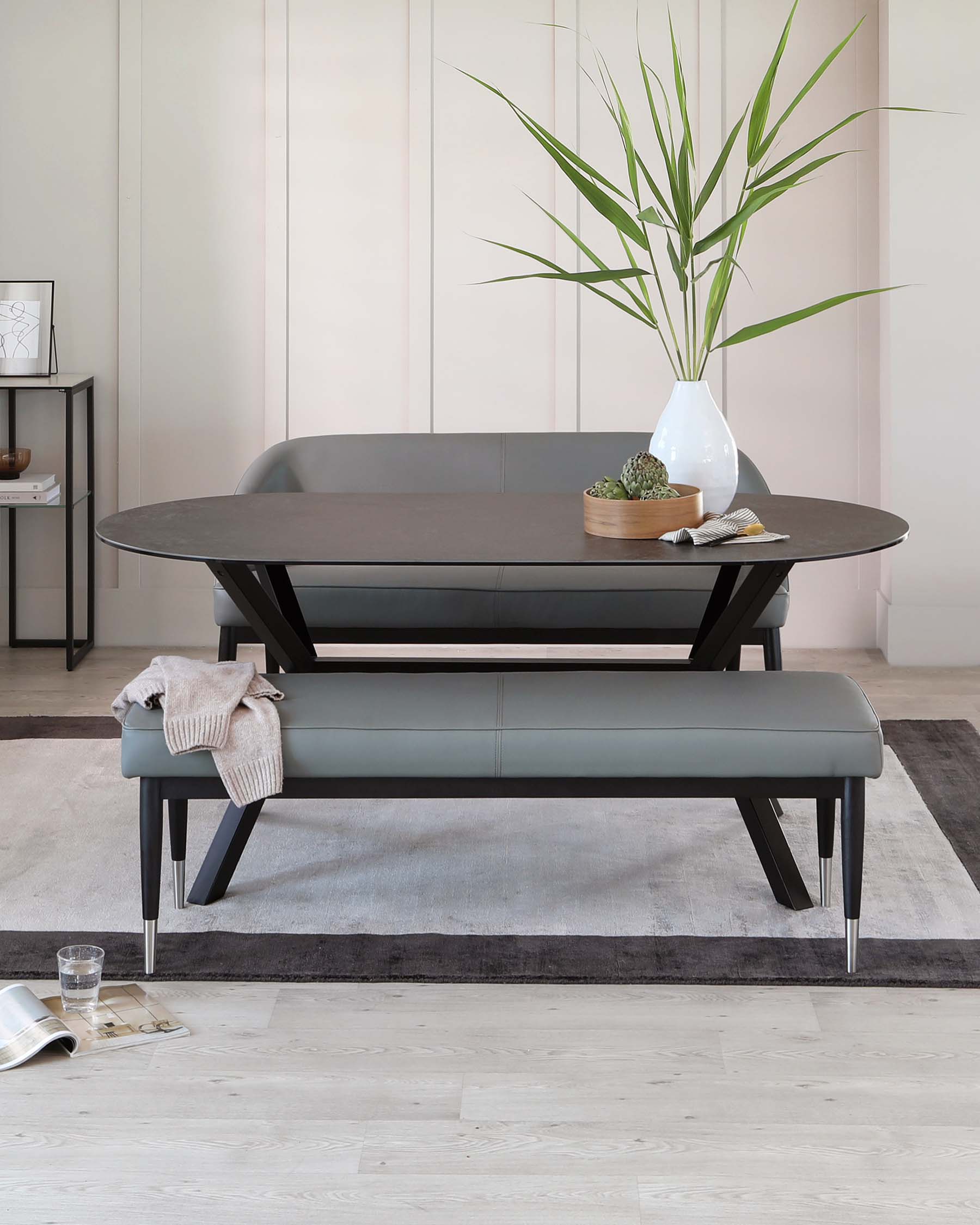 Modern dark wood coffee table with rounded corners and sleek metal legs, paired with a matching minimalist leather bench featuring clean lines and metal accents. The table is styled with a white vase containing green plants and a small wooden tray with succulents, accompanied by a casual drape of a light beige sweater over the bench. A black and white magazine and a water glass rest on a light wood floor beside a dark grey area rug.