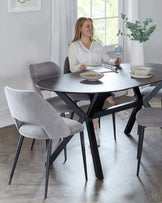 Modern dining set featuring a round, dark grey tabletop with black angular legs, accompanied by three light grey upholstered chairs with a curved backrest and tapered black metal legs. A white ceramic vase with greenery adds a decorative touch on the table.