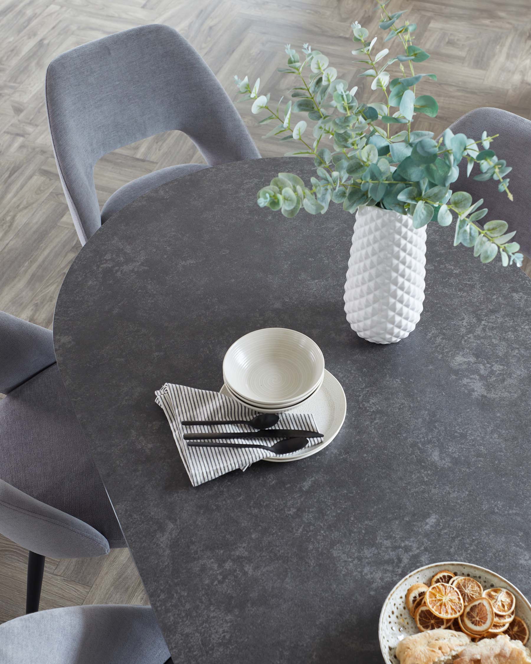 Round, modern dining table with a stone-like matte finish and three cushioned grey fabric chairs with a curved backrest.